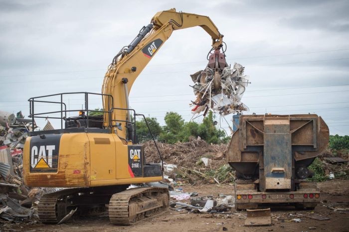 An excavator with a claw attachment loads light-gauge steel into a machine that will compress it into bricks.