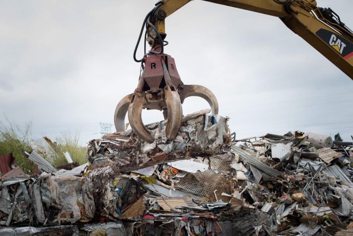 An excavator fitted with a claw loads a bale of recycled steel onto a stack of other bales.