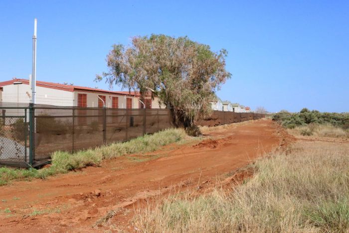 Dongas behind a fence next to a red dirt road and grass.