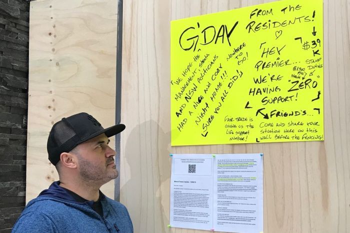 A man in a hat stands in front of a yellow sign stuck on a wall.