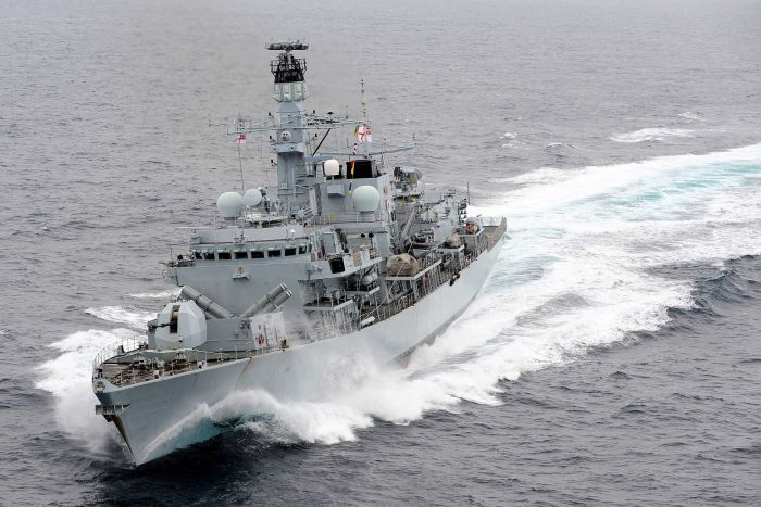 A grey Royal Navy ship cuts through grey sea waters on an overcast day. 