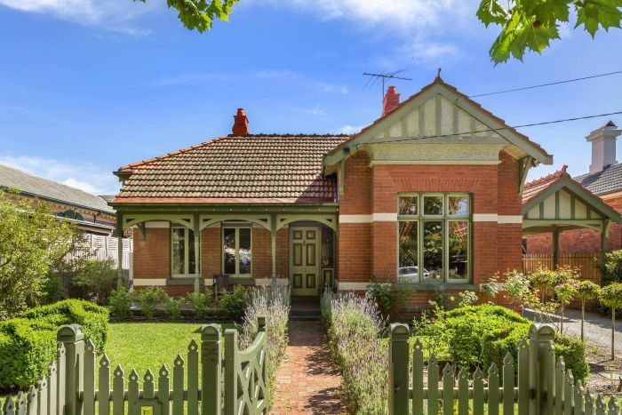 A historic double-fronted brick house sits behind a green picket fence and manicured front garden