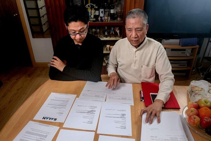 Man with glasses and black shirt next to older man in cream shirt, sitting at dining table with papers scattered in front.