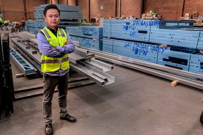 Man wearing high-vis vest and blue collared shirt, standing arms crossed in a warehouse with large steel behind him.