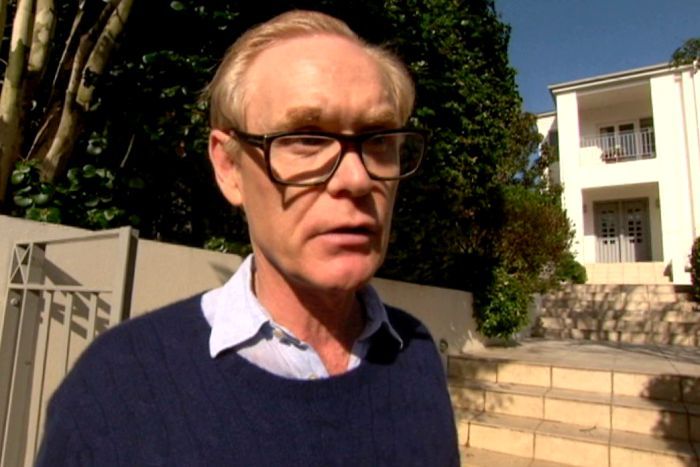 Man with glasses and blue jumper stands outside large home with gate and trees behind him.